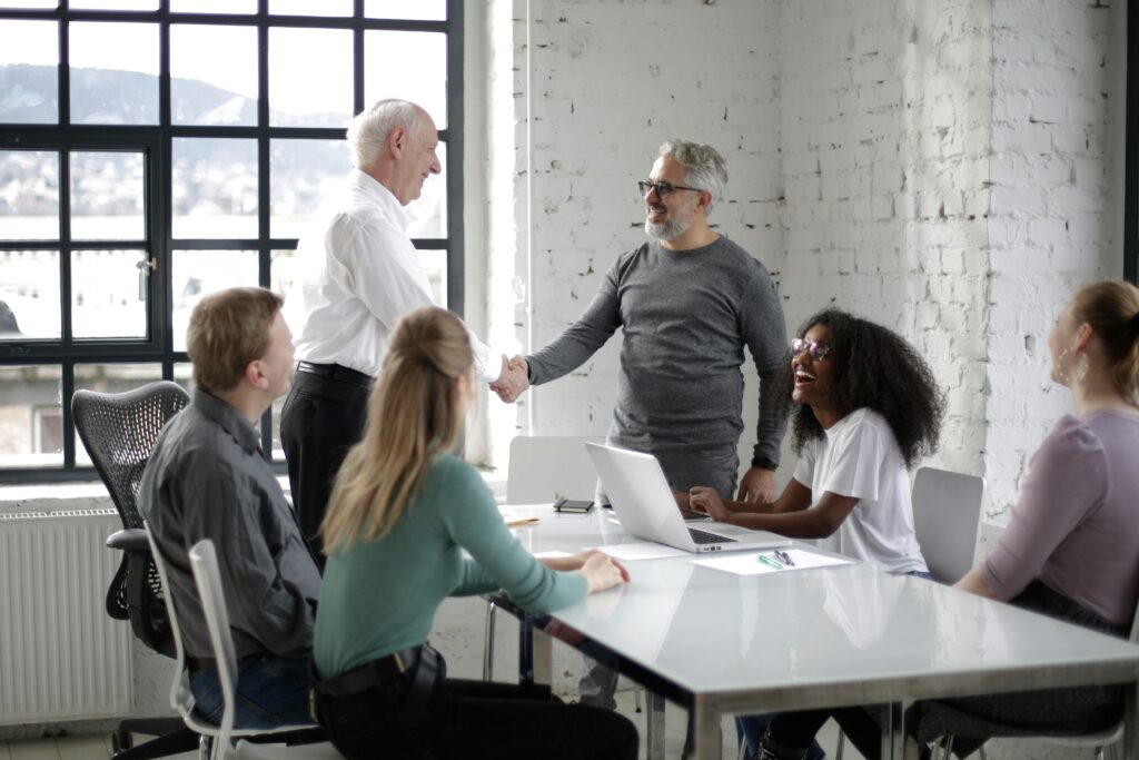 Cheerful male colleagues shaking hands while discussing business ideas with group of multiethnic coworkers gathering around table with gadgets and documents in modern light workspace